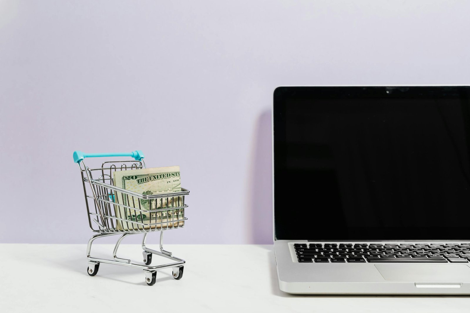 Mini shopping cart with cash beside a laptop, symbolizing online shopping and e-commerce.